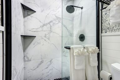Spa-like modern bathroom: marble tile shower with glass enclosure, black matte rain showerhead and corner shelves, white towels folded on a towel bar and patterned wall accent.
