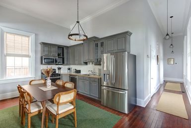 Bright open-plan kitchen and dining area with gray shaker cabinets, stainless steel French-door fridge, wooden dining table with cane-back chairs on a green rug, hardwood floors, large window, and pendant chandelier lighting along a long hallway.