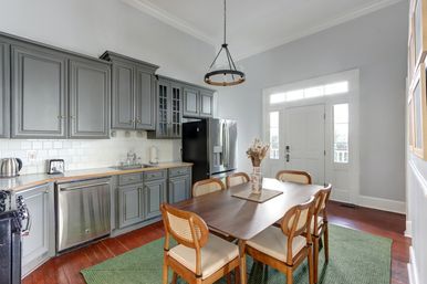 Bright transitional kitchen-dining area with gray shaker cabinets, stainless steel refrigerator and dishwasher, wooden dining table with cane-back chairs, round pendant light, green area rug, hardwood floors and white front door.