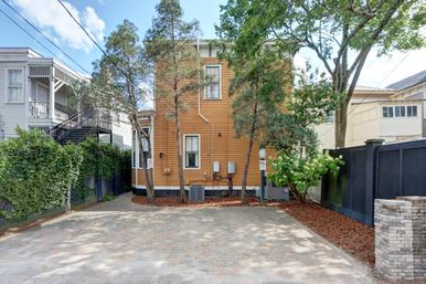 Sunny rear view of a two-story yellow wooden house with mature trees, utility meters and an AC unit, bordered by a black privacy fence and ivy-covered wall, opening onto a paved brick parking pad in an urban residential alley.