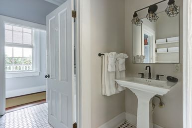 Bright airy residential bathroom with white pedestal sink and matte-black faucet, industrial cage vanity lights, folded white towels on a black towel bar, hexagon tile floor and open door to a sunlit room with large window.