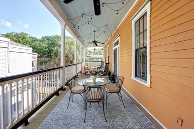 Covered second-floor porch with orange siding, hanging string lights and ceiling fans, glass-top table with four metal chairs on a patterned rug, rocking chairs and potted plants overlooking a tree-lined neighborhood.
