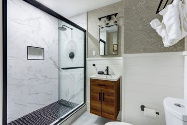 Spa-like modern bathroom with glass-enclosed marble-look shower, black hex-tile shower floor and matte-black fixtures, floating wood vanity, mirror, white shiplap walls and folded towels