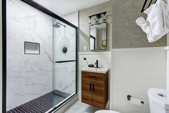 Spa-like modern bathroom with glass-enclosed marble-look shower, black hex-tile shower floor and matte-black fixtures, floating wood vanity, mirror, white shiplap walls and folded towels