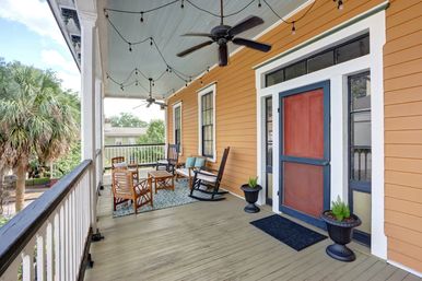 Sunny southern-style covered front porch with orange siding and red door, seating area with rocking chairs and wooden chairs on a patterned rug, ceiling fans and string lights, potted plants, and palm-tree view.