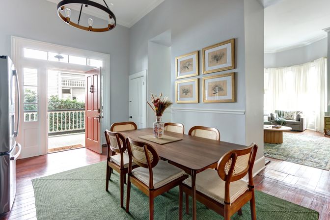Welcoming sunlit dining room with wooden table and six cane-back chairs on a green rug, red front door open to a porch, framed botanical prints on the wall, and an adjacent bay-window living room visible.