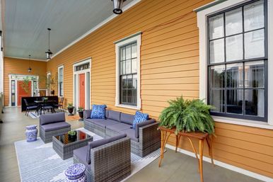 Bright covered porch with orange clapboard siding, gray wicker sofa and chairs with blue pillows, glass-top coffee table, potted fern on wooden stand and outdoor dining area