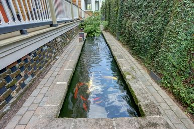 Narrow rectangular koi pond with colorful koi gliding between brick pavers, an ivy-covered wall, and a wooden porch railing in a shaded courtyard.