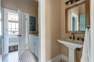 Cozy home bathroom with white pedestal sink, matte black faucet and framed mirror, hexagon tile floor, and an open door revealing a side-by-side washer and dryer beneath a sunlit window with a bamboo shade.