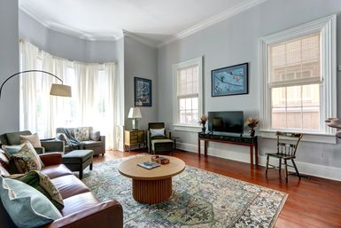 Sunlit townhouse living room with bay window, hardwood floors, patterned rug, leather sofa, cozy armchairs, round wooden coffee table and TV console.