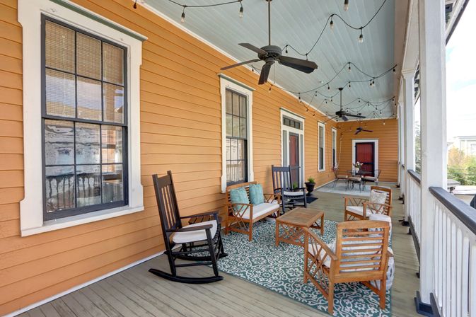 Long covered front porch with orange clapboard siding and pale blue ceiling, furnished with wooden rocking chairs, cushioned patio seating, patterned outdoor rug, ceiling fans, string lights, and a small coffee table.