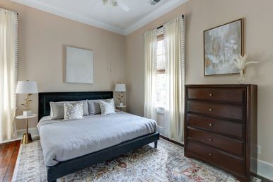 Cozy sunlit bedroom with gray upholstered bed, patterned area rug, dark wood five-drawer dresser, sheer floor-to-ceiling curtains over tall windows, brass bedside lamps, decorative throw pillows and abstract wall art.