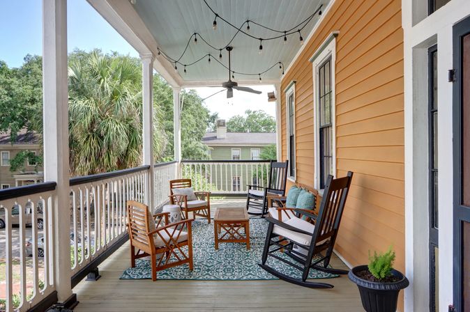 Sunny, inviting southern-style front porch with warm orange siding, string lights and ceiling fan, patterned rug, wooden chairs and rocking chairs around a coffee table, potted fern and palm-tree-lined neighborhood view.