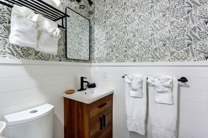 Cozy modern bathroom interior with botanical floral wallpaper, white shiplap wainscoting, wood vanity with black faucet and mirror, plus neatly folded white towels on black racks.