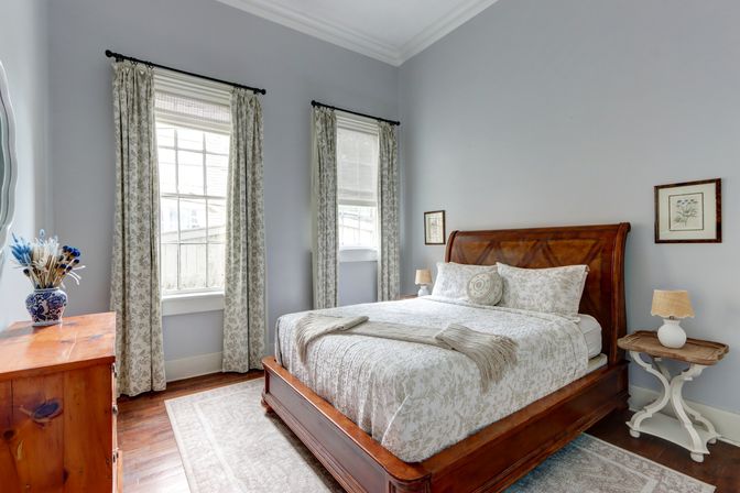 Cozy sunlit bedroom with a wooden sleigh bed and soft floral bedding, light gray walls, two tall windows with patterned curtains, hardwood floors, wooden dresser with blue vase, and bedside tables with lamps.