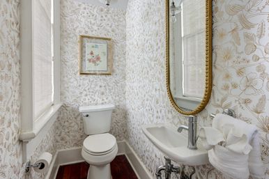 Cozy small powder room with white toilet and wall-mounted sink, vintage gold-framed oval mirror, beige floral wallpaper, hardwood floor, and a window with blinds.