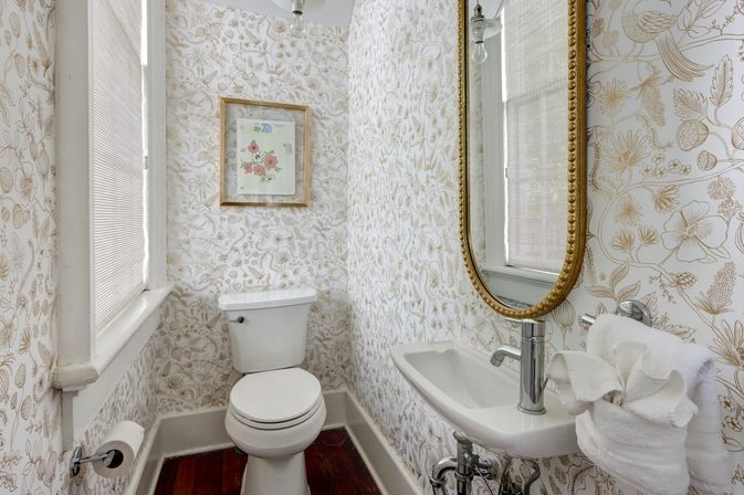 Cozy small powder room with white toilet and wall-mounted sink, vintage gold-framed oval mirror, beige floral wallpaper, hardwood floor, and a window with blinds.