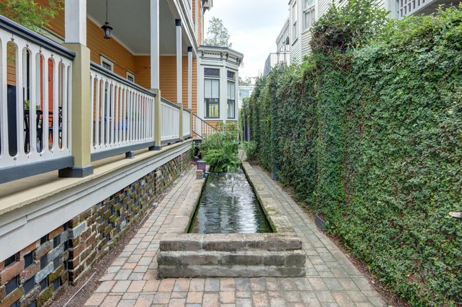 Charming narrow courtyard with a long rectangular reflecting pool, brick pavers, a yellow Victorian-style porch with white railings on the left and a tall ivy-covered wall on the right.