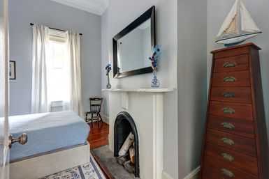 Bright coastal-inspired bedroom with bed, white mantel fireplace topped by a large mirror, tall wooden drawer chest with sailboat model, hardwood floors and a sunlit window with sheer curtains.