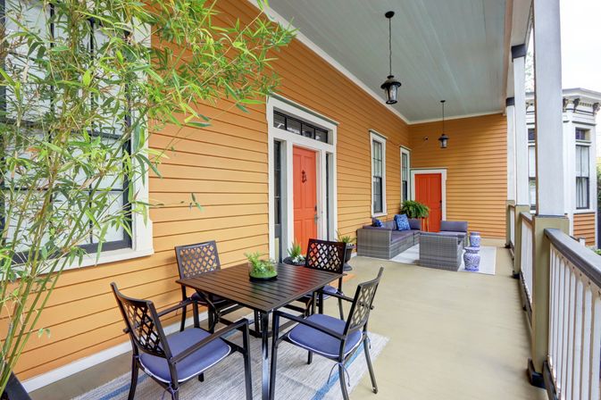 Inviting covered front porch with orange siding and red doors, outdoor dining table, cushioned wicker lounge seating, hanging lanterns and potted plants.