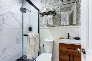 Bright modern bathroom with glass-enclosed shower featuring marble-look tile and black rain showerhead, white toilet, wood vanity with black faucet, patterned wallpaper and neatly folded white towels