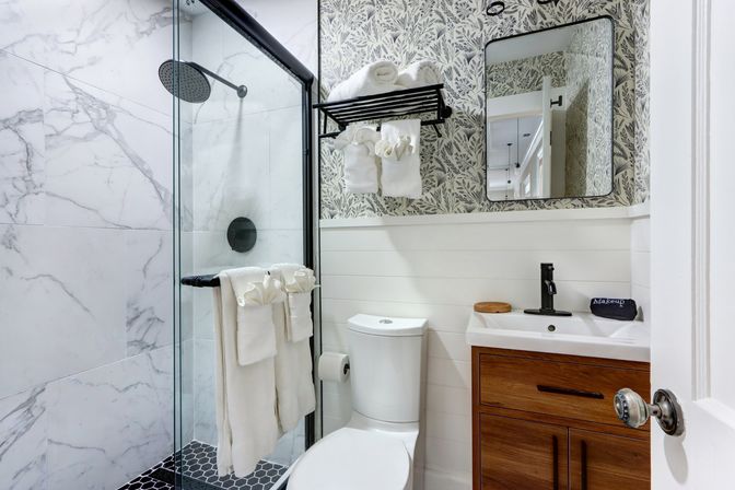 Bright modern bathroom with glass-enclosed shower featuring marble-look tile and black rain showerhead, white toilet, wood vanity with black faucet, patterned wallpaper and neatly folded white towels