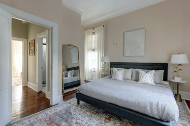 Contemporary light-filled neutral master bedroom with a dark upholstered king bed, patterned area rug, hardwood floors, full-length gold mirror, sheer curtains, bedside tables with lamps, and minimalist wall art.