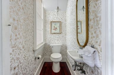 Cozy powder room with beige floral wallpaper, white toilet, wall-mounted sink topped by a gold oval mirror, framed floral print, window blind, and dark hardwood floor.