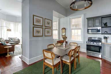 Sunlit open-plan dining and kitchen with a mid-century wooden table and six cane-back chairs on a green rug, gray cabinets and stainless-steel range, framed tree prints on pale walls, and a cozy living room visible through the doorway.