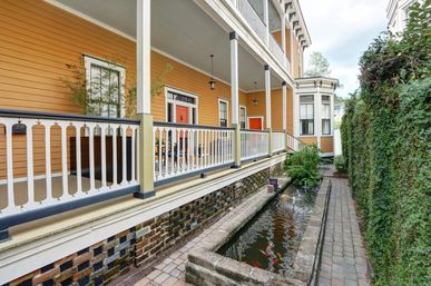 Charming yellow Victorian-style two-story home with covered porch and white decorative railing, red doors, narrow koi pond with colorful fish beside a brick walkway and ivy-covered wall.