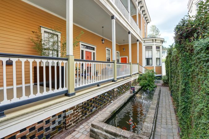 Charming yellow Victorian-style two-story home with covered porch and white decorative railing, red doors, narrow koi pond with colorful fish beside a brick walkway and ivy-covered wall.