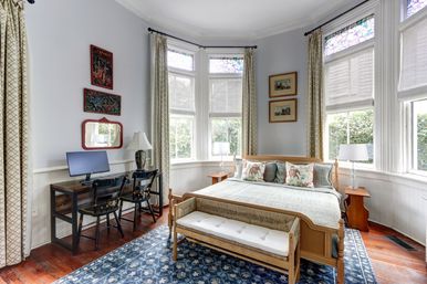 Sunlit Victorian-style bedroom with bay windows and stained-glass transoms, wooden bed with patterned pillows and cushioned bench, blue floral rug, side tables and lamps, and a small wooden desk with computer and chairs.