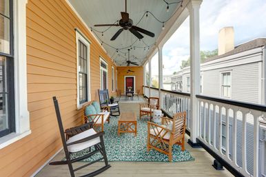 Cozy second-floor Victorian-style porch with orange clapboard siding, white balustrade, teal patterned rug, wooden chairs and rocking chairs, ceiling fans and string lights overlooking neighboring homes.