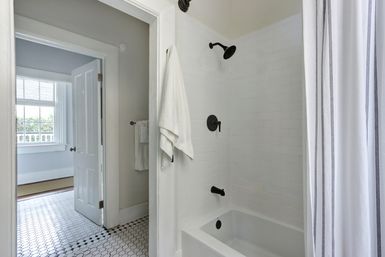 Bright white bathroom with subway-tiled shower and bathtub, matte black fixtures, striped shower curtain, white towels and hexagon tile floor, open door to a sunlit room with large window.