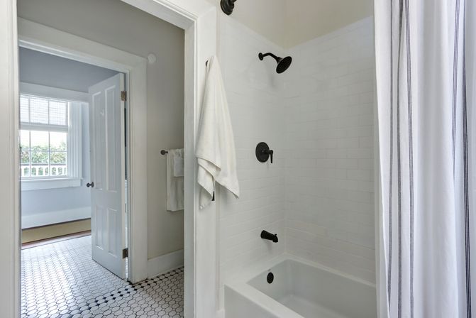 Bright white bathroom with subway-tiled shower and bathtub, matte black fixtures, striped shower curtain, white towels and hexagon tile floor, open door to a sunlit room with large window.