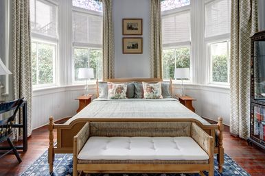 Cozy sunlit bedroom with wooden bed, tufted bench, decorative pillows, bay windows with stained-glass transoms, matching lamps, patterned curtains and hardwood floors.