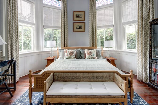 Cozy sunlit bedroom with wooden bed, tufted bench, decorative pillows, bay windows with stained-glass transoms, matching lamps, patterned curtains and hardwood floors.