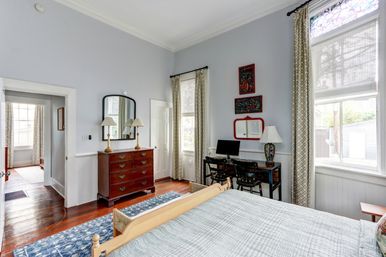 Sunlit bedroom in a historic home — high ceilings, crown molding, hardwood floors, blue patterned rug, wooden bed with quilt, dresser with mirror and lamps, small desk with computer and patterned curtains.