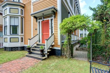 Yellow Victorian-style house exterior with a coral front door, porch steps flanked by black urn planters, a brick walkway, and a leafy arbor opening to a narrow side courtyard with a water feature.