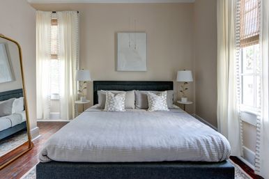 Bright, cozy neutral master bedroom interior with a gray upholstered king bed, cream linen bedding and patterned throw pillows, matching round bedside tables and lamps, tall cream curtains over hardwood floors, and a large leaning mirror.