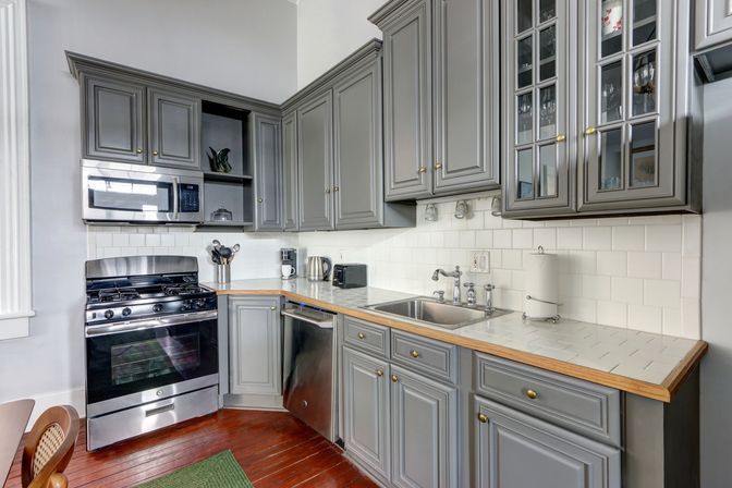 Cozy kitchen with gray cabinets, stainless-steel gas range, microwave and dishwasher, white subway-tile backsplash, tiled countertops with wood trim and hardwood floors.