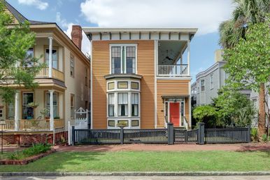 Historic Victorian-style two-story townhouse with mustard-orange siding, decorative bay window, covered second-floor porch, red front door, gray picket fence, palm tree and neighboring homes under a blue sky.