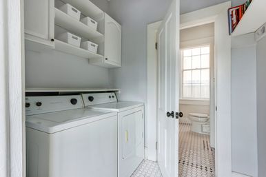 Bright residential laundry room with white top-load washer and matching dryer under open shelving with baskets and cabinets, open door to a sunlit bathroom with a hexagon-tile floor.