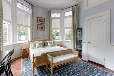 Sunlit bay-window bedroom with stained-glass transoms, light wood bed and bench, floral throw pillows, patterned blue area rug, bedside tables and lamps