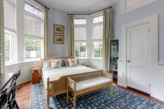 Sunlit bay-window bedroom with stained-glass transoms, light wood bed and bench, floral throw pillows, patterned blue area rug, bedside tables and lamps
