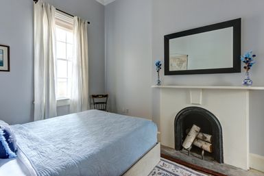 Sunlit, airy bedroom with light-blue quilted bed, tall window with sheer curtains, small vintage chair, white mantel with a black arched fireplace holding birch logs, large rectangular mirror and blue floral vases.