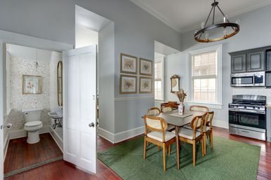Bright dining area with wooden table and rattan chairs on a green rug, open to a compact bathroom and a modern stainless-steel kitchen, hardwood floors and soft gray walls with abundant natural light.