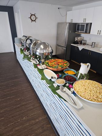 Indoor catering buffet in a bright kitchen: three stainless-steel chafing dishes on a striped counter with bowls of mixed salad, pasta salad, toppings and dressings.