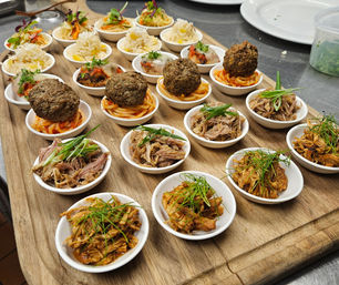 Assorted bite-sized appetizers on a wooden serving board: mini meatballs on spaghetti, pulled pork and shredded chicken in small white bowls, each garnished with fresh herbs.