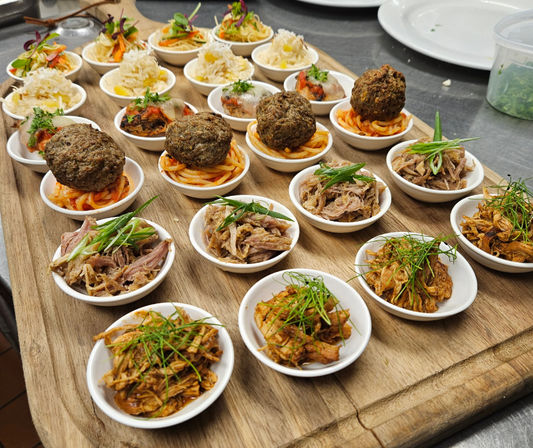 Assorted bite-sized appetizers on a wooden serving board: mini meatballs on spaghetti, pulled pork and shredded chicken in small white bowls, each garnished with fresh herbs.